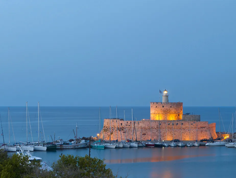 Mandraki Harbor in Rhodes at dusk with ancient fortress walls and boats docked in the calm water.