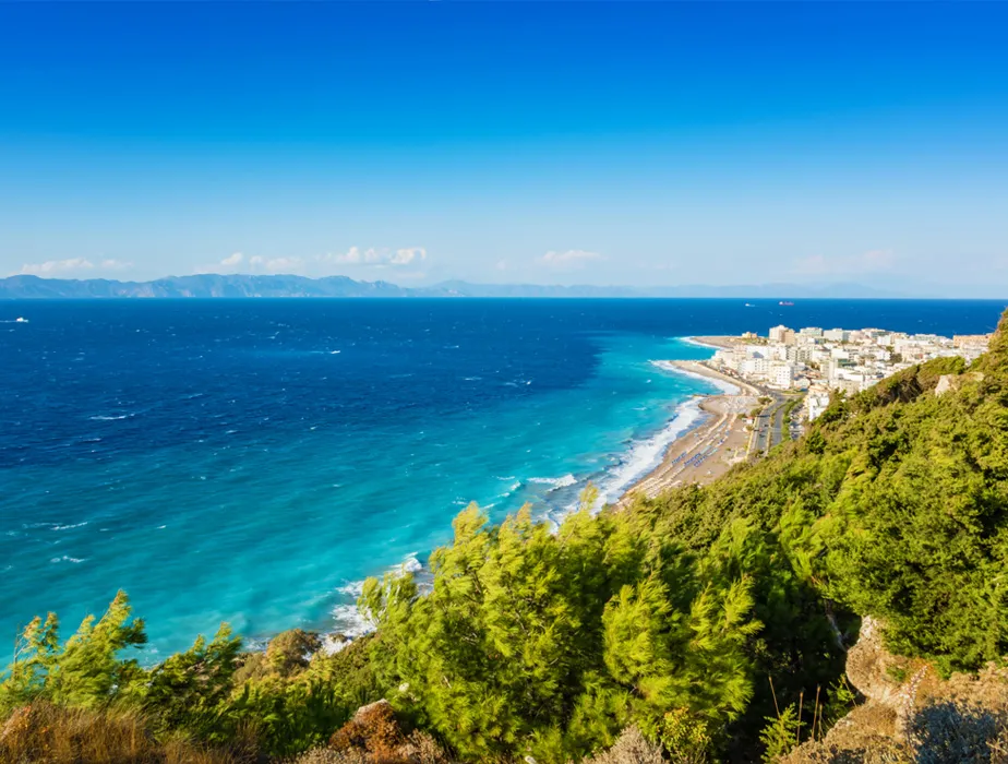 Coastal view of Rhodes, Greece, with the town, beach, turquoise sea, and mountains in the distance.