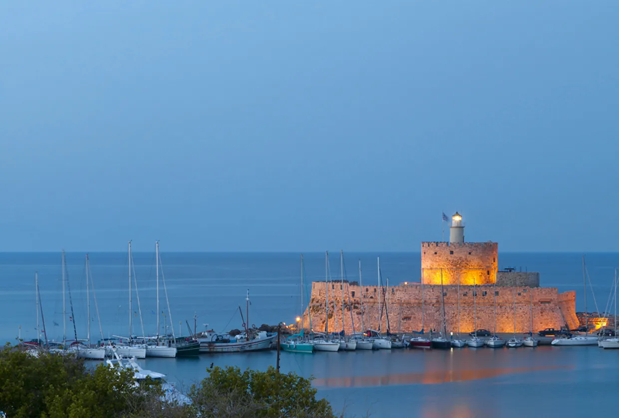 Porto di Mandraki a Rodi al crepuscolo con barche a vela attraccate e fortezza con faro illuminato sul mare.