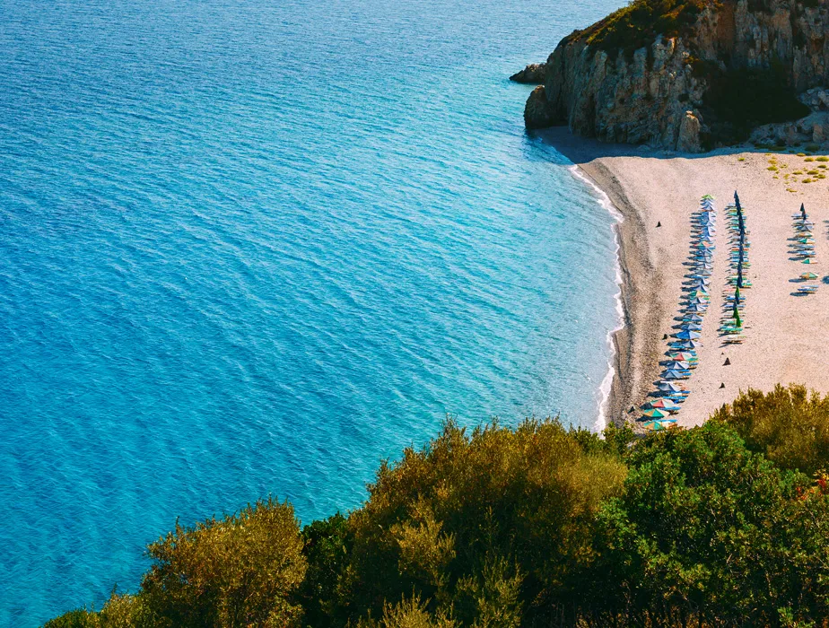 Bella costa con spiaggia di ciottoli e ombrelloni organizzati accanto a acqua limpida e scogliere rocciose.