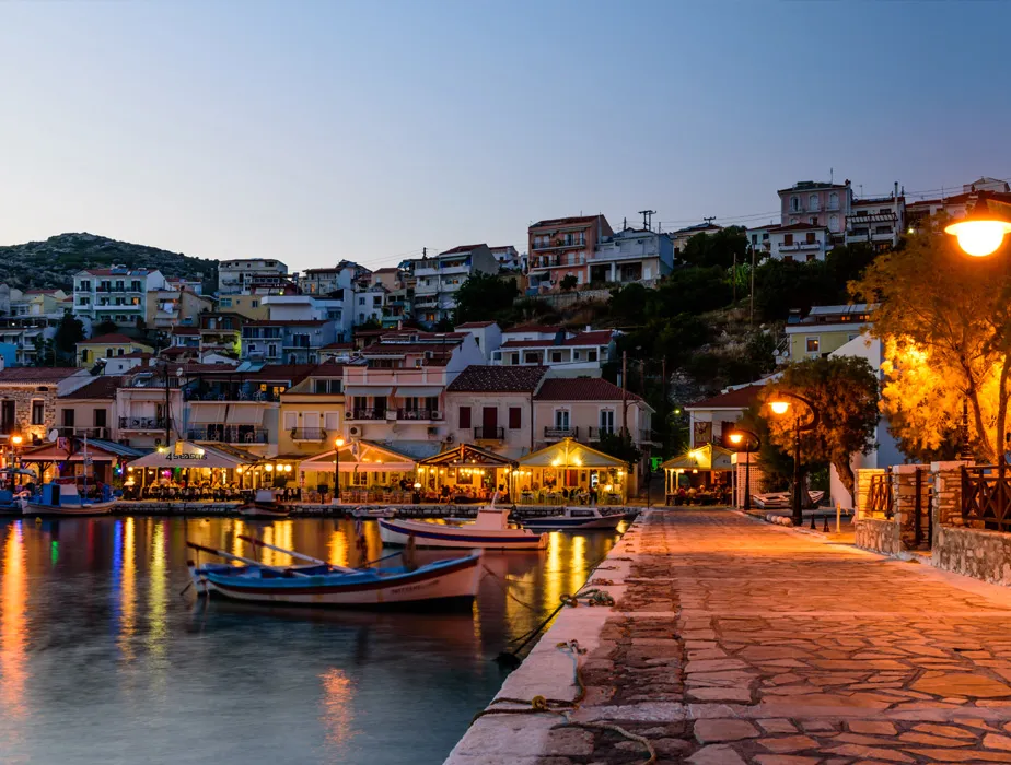 Veduta del porto di Parga al crepuscolo con taverne illuminate e barche ancorate per auto a noleggio.