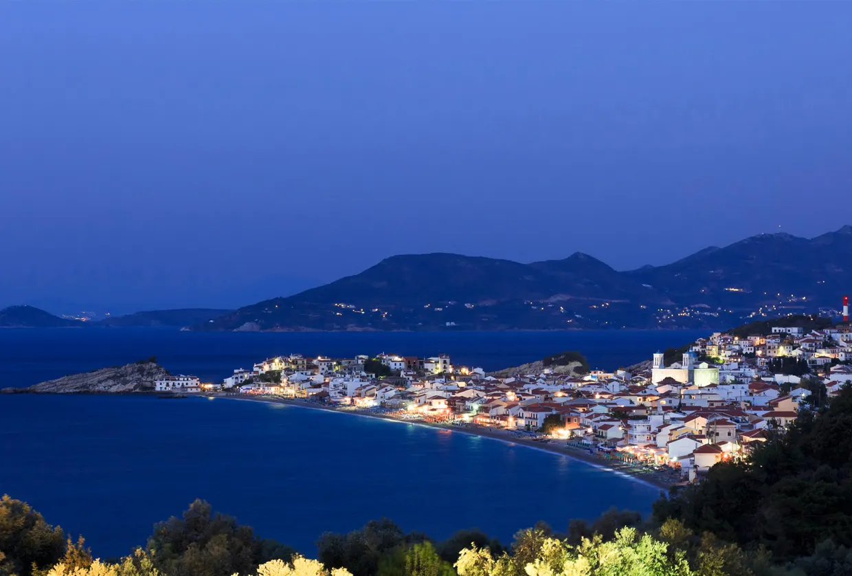 Volos coastal town illuminated at dusk with a beach and mountains in the background for car rental trips.