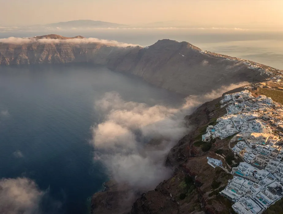 Santorini island coastline with white buildings and cliffs above a cloudy sea at sunset.