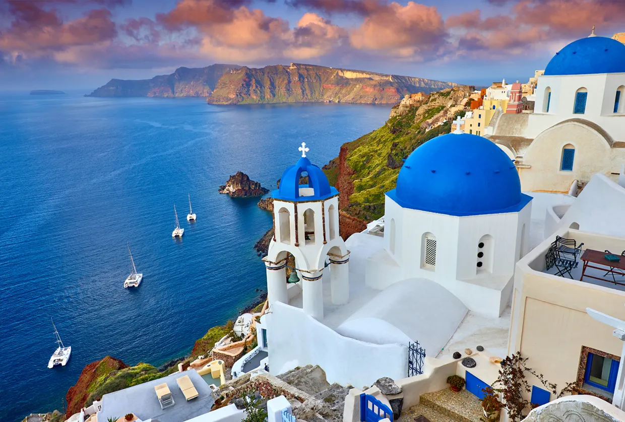 Santorini coastline with blue-domed churches and sailboats in the sea during sunset light.