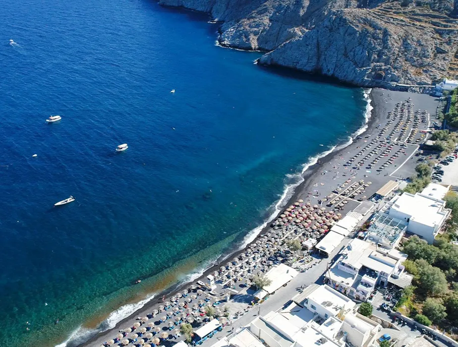 Santorini beach with sun umbrellas and boats in the deep blue sea by rocky cliffs.