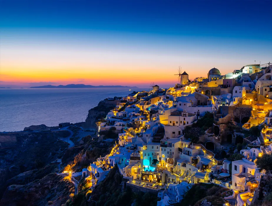 Côte de Santorin au coucher du soleil avec bâtiments blancs et moulins éclairés sur les falaises.