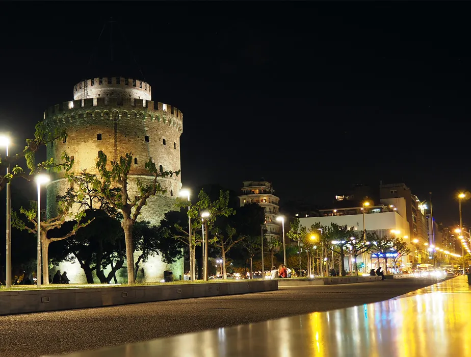 White Tower of Thessaloniki lit up at night along the waterfront promenade with streetlights and trees.