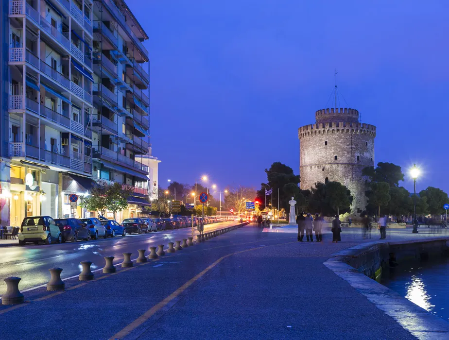 White Tower of Thessaloniki at dusk with people near the waterfront and parked cars along the street.
