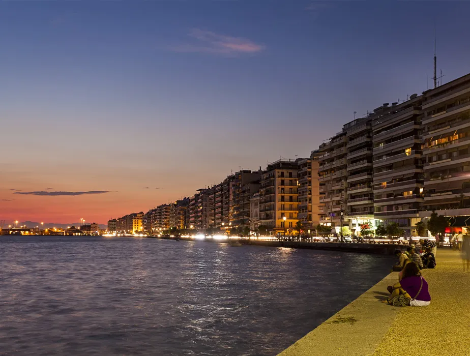 Thessaloniki waterfront at sunset with buildings, lights, and people relaxing by the sea.