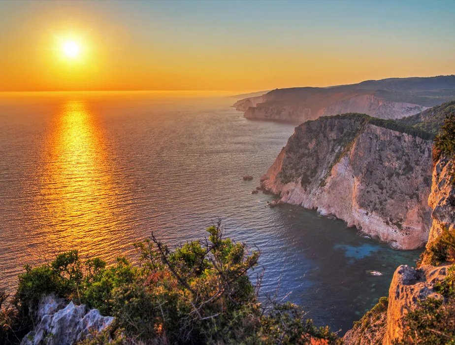 Tramonto su una costa rocciosa con acqua limpida e vegetazione, ideale per noleggio auto lungo la costa.