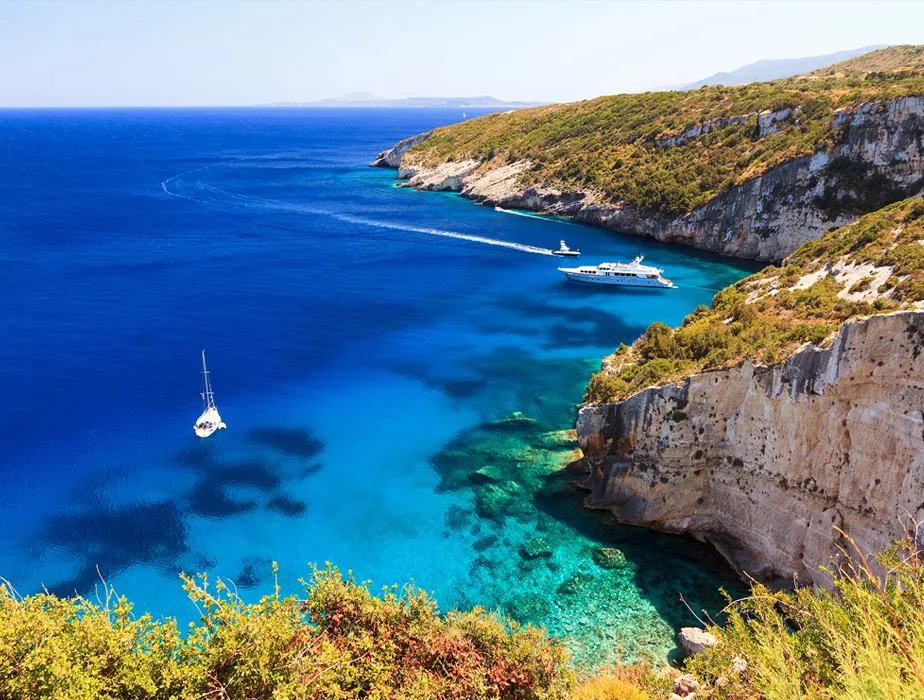 Costa mediterranea con acqua limpida e blu e barche vicino a scogliere rocciose e vegetazione.