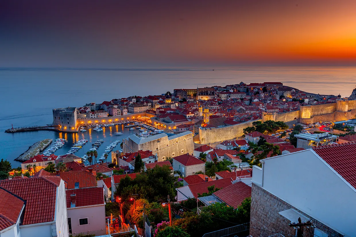 Dubrovnik old town at sunset with illuminated city walls and boats in the harbor by the Adriatic Sea.