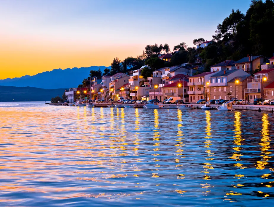 Coastal town at sunset with waterfront houses and boats reflecting on calm water.