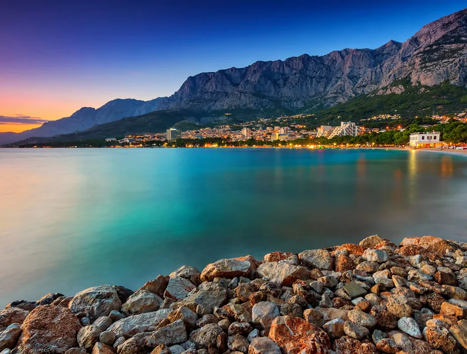 Makarska coastline with rocky shore, calm turquoise sea and mountain backdrop at sunset in Croatia.