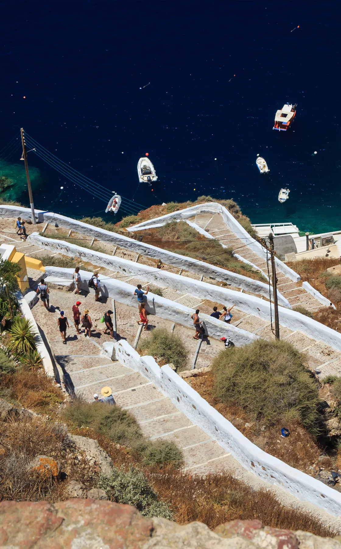 Santorini coastal view with tourists walking down white stone stairs near blue sea and boats for rent.