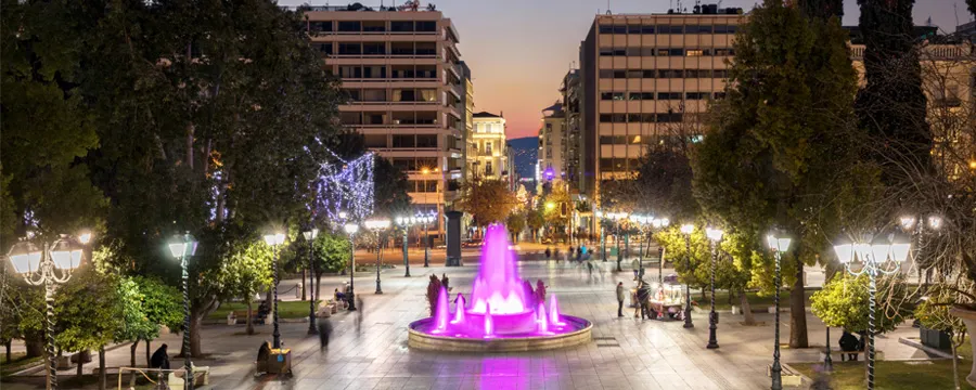 Syntagma Square in Athens at dusk with illuminated pink fountain and people walking along the pedestrian street.
