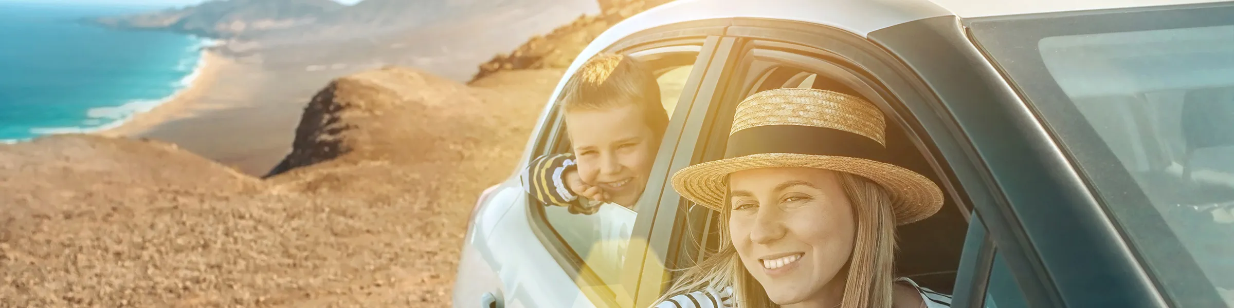 Donna sorridente con cappello di paglia e bambino che guardano dalle finestre di un'auto a noleggio in costa.
