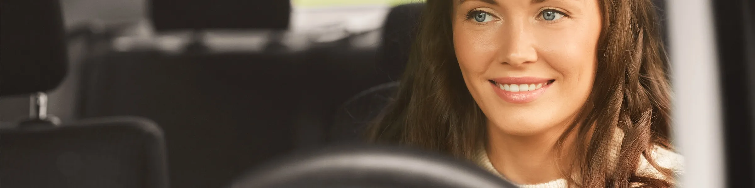 Smiling woman with blue eyes and wavy brown hair driving a car, focusing on road safety and comfort.