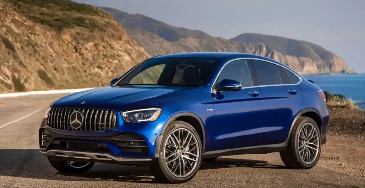 Ενοικίαση πολυτελούς αυτοκινήτου Mercedes-Benz GLC Coupe με κομψό μπλε σχεδιασμό κοντά σε παραλιακό δρόμο και λόφους.