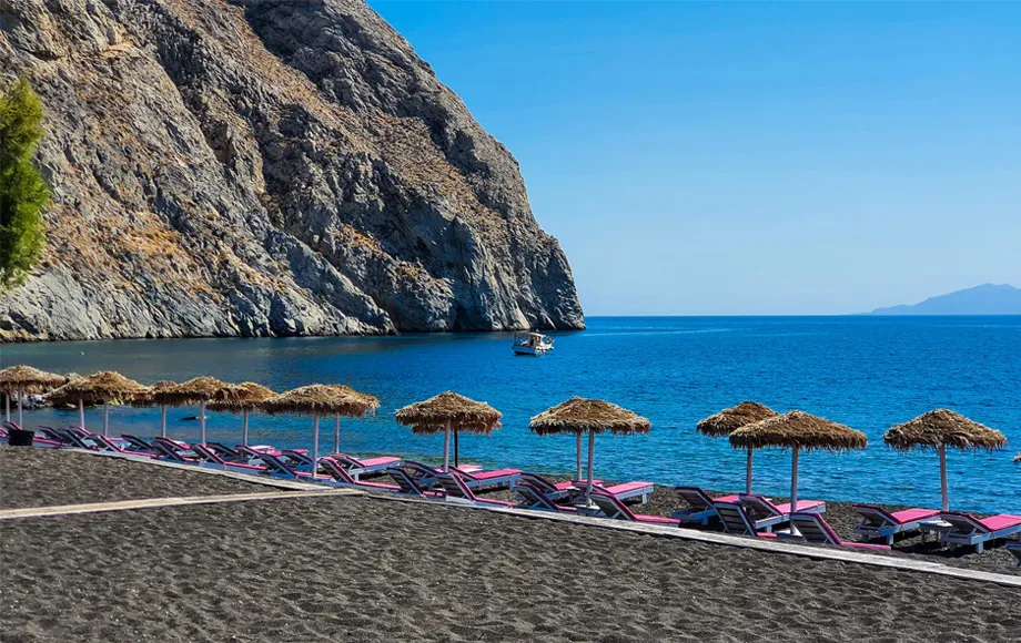 Black sand beach in Santorini with sun loungers and straw umbrellas by the calm blue sea.