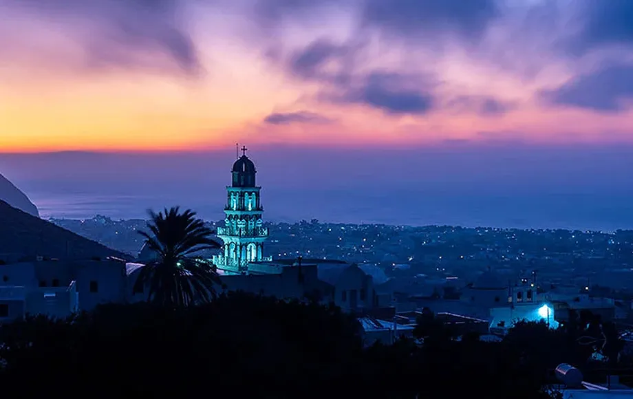 Sunset view over Santorini with illuminated church tower and town lights at dusk.