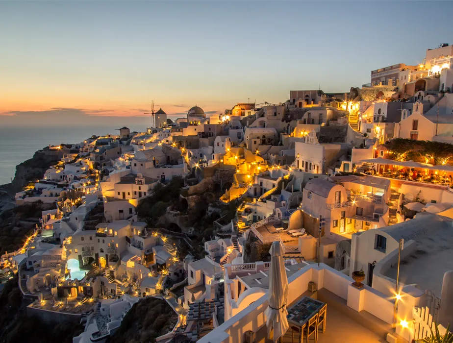 Santorini at sunset with white buildings and glowing lights on the cliffs, perfect for car rental travel.