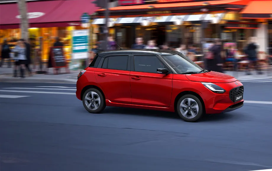 Mașină de închiriat Suzuki Swift roșie care circulă pe o stradă din oraș cu pietoni neclari în fundal.